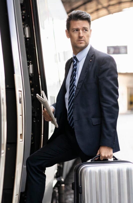 A man in a suit, holding a newspaper and a suitcase, steps onto a train.